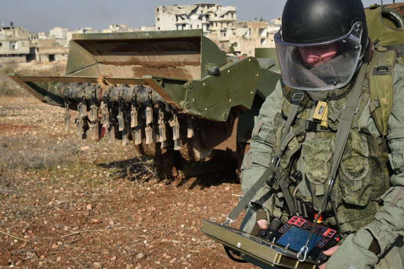 A Russian soldier concentrates on his handheld computer as the buildings of Aleppo stand crumbling behind him and his military digger